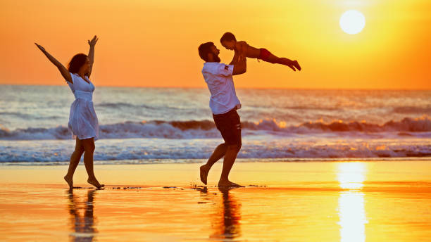 Happy family walking with fun on sunset sea beach Happy family - father, mother, baby son walk with fun along edge of sunset sea surf on black sand beach. Active parents and people outdoor activity on summer vacations with children on Bali island raro stock pictures, royalty-free photos & images