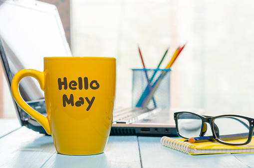 Hola mayo - texto en taza de café amarillo en el fondo de la oficina de negocios, lugar de trabajo con ordenador portátil y gafas. Tiempo de primavera, espacio vacío para el texto photo