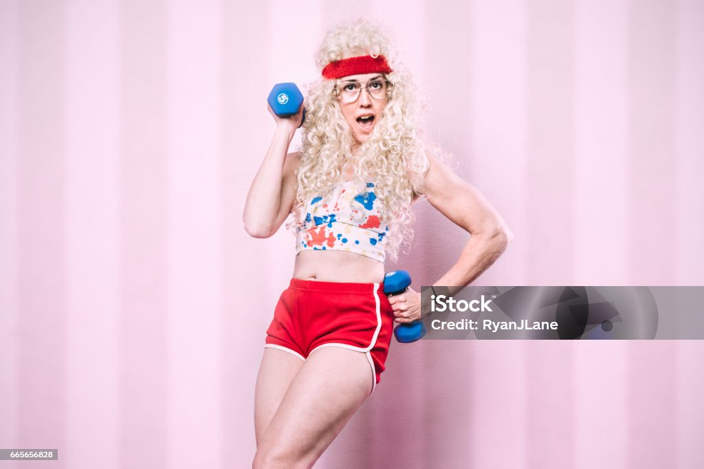 Aerobics Teacher Working Out An aerobics class instructor from the late 1980's - early 1990's poses for a portrait, her long blond hair styled with a big perm.  She exercises with small dumbbells, getting a good cardio workout in.  Pink striped background. Humor Stock Photo