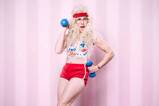 An aerobics class instructor from the late 1980's - early 1990's poses for a portrait, her long blond hair styled with a big perm.  She exercises with small dumbbells, getting a good cardio workout in.  Pink striped background.