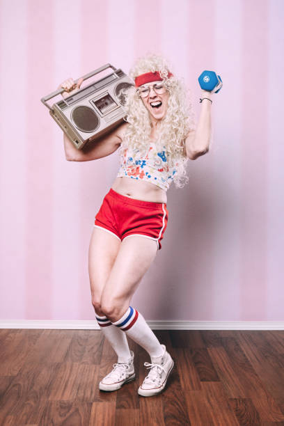 Aerobics Teacher Working Out An aerobics class instructor from the late 1980's - early 1990's poses for a portrait, her long blond hair styled with a big perm.  She exercises with small dumbbells, getting a good cardio workout in while holding her stereo boombox.  Pink striped background. 80s aerobics stock pictures, royalty-free photos & images