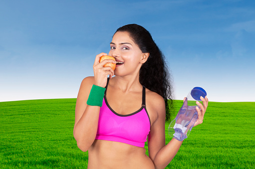 Portrait of young woman eating an apple  while holding a bottle in the meadow