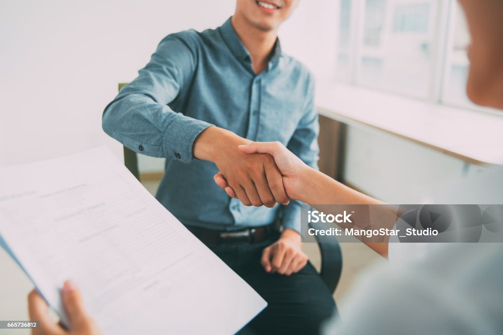 Smiling Asian businessman shaking partners hand Smiling Asian businessman wearing shirt sitting in office and shaking hand of female partner. Woman holding document Job Interview Stock Photo