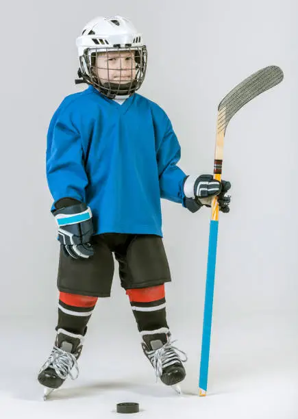 Cute little boy in ice-hockey uniform and in skates is standing holding hockey stick in his hand. The boy hockey player is looking menacing at the camera. The puck lying on the floor in front of him. Studio shooting on a white background