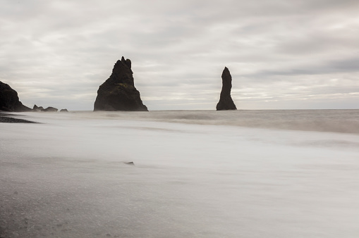 Scenic view of Reynisdrangar basalt sea stacks in sea. Beautiful view of black sand beach against cloudy sky. It is one of the famous tourist attraction in Iceland.