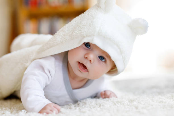 Baby girl wearing white towel or winter overal in white sunny bedroom Baby girl with blue eyes wearing white towel or winter overal in white sunny bedroom. Newborn child relaxing in bed. Nursery for children. Textile and bedding for kids. New born kid with toy bear. baby boys stock pictures, royalty-free photos & images