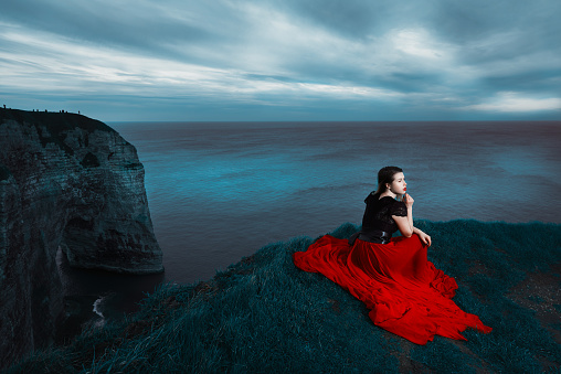 dramatic and nostalgic woman sitting at the edge of cliff, looking away and thinking. wearing red dress.