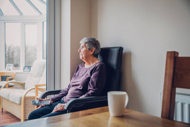 Senior Woman Sitting Alone Senior woman sitting alone in her kitchen. She is looking out into her conservatory while holding a home telephone in her hand. Serious expression neighborhood crime watch stock pictures, royalty-free photos & images
