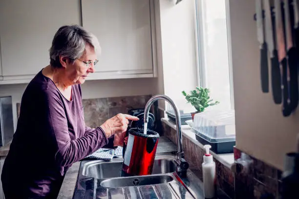 Elderly woman filling up a kettle while standing at her kitchen sink.