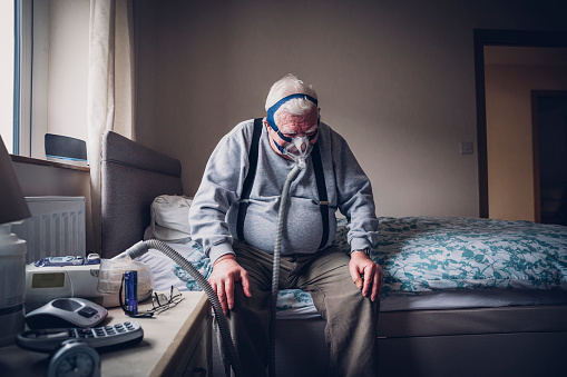 Elderly man sitting at home on his bed. He is wearing a medical breathing apparatus over his face attached to a machine beside him.