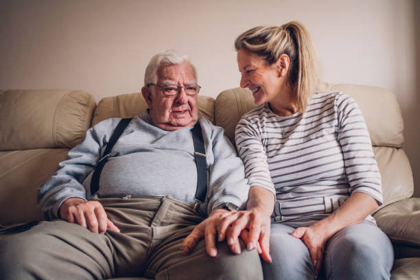 Senior Man Relaxing with his Daughter The senior man sits on the sofa in his living room beside his daughter. The woman smiles at her father with her hand resting on his. father daughter 80s stock pictures, royalty-free photos & images