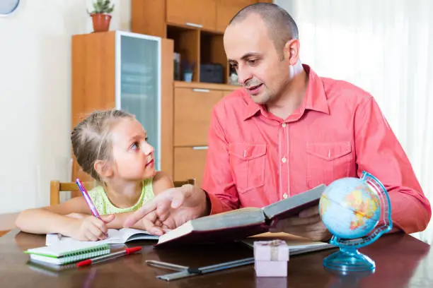 Adult male tutor and little girl studying with books in domestic interior