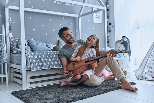 Young father teaching his little daughter to play guitar and smiling while sitting on the floor in bedroom