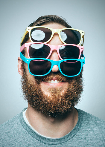 A portrait of a man with a large beard, wearing multiple pairs of multi-colored sunglasses.  Studio shot.