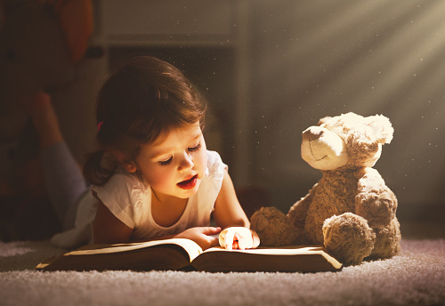 Little child girl is reading a book in evening in dark with a toy bear