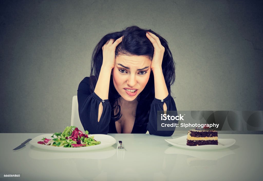 Woman tired of diet restrictions Woman tired of diet restrictions deciding whether to eat healthy food or sweet cake dessert she is craving isolated grey background. Human face expression emotion. Food Stock Photo