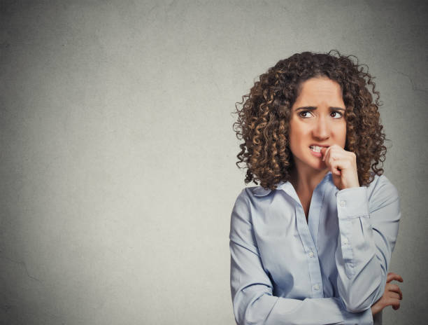 unpleased skeptical young curly brown hair woman Closeup portrait nervous looking woman biting her fingernails craving something anxious isolated grey wall background with copy space. Negative human emotion facial expression body language perception Blush stock pictures, royalty-free photos & images