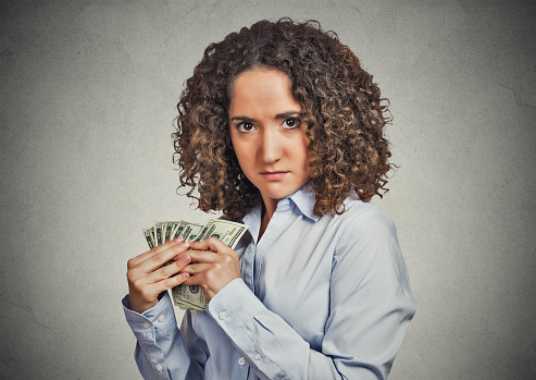 Closeup portrait greedy young woman corporate business employee, worker, student holding dollar banknotes tightly isolated grey wall background. Negative human emotion facial expression feeling