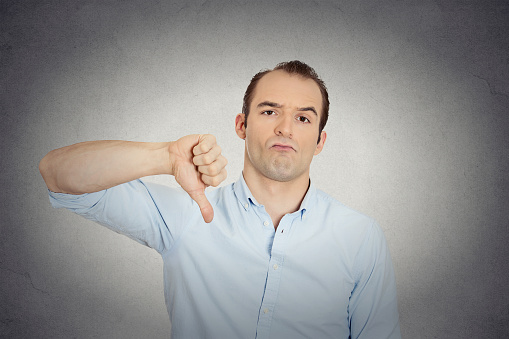 Closeup portrait of angry, unhappy, young handsome man showing thumbs down sign, in disapproval of offer, situation isolated grey wall background. Negative human emotion facial expression feelings