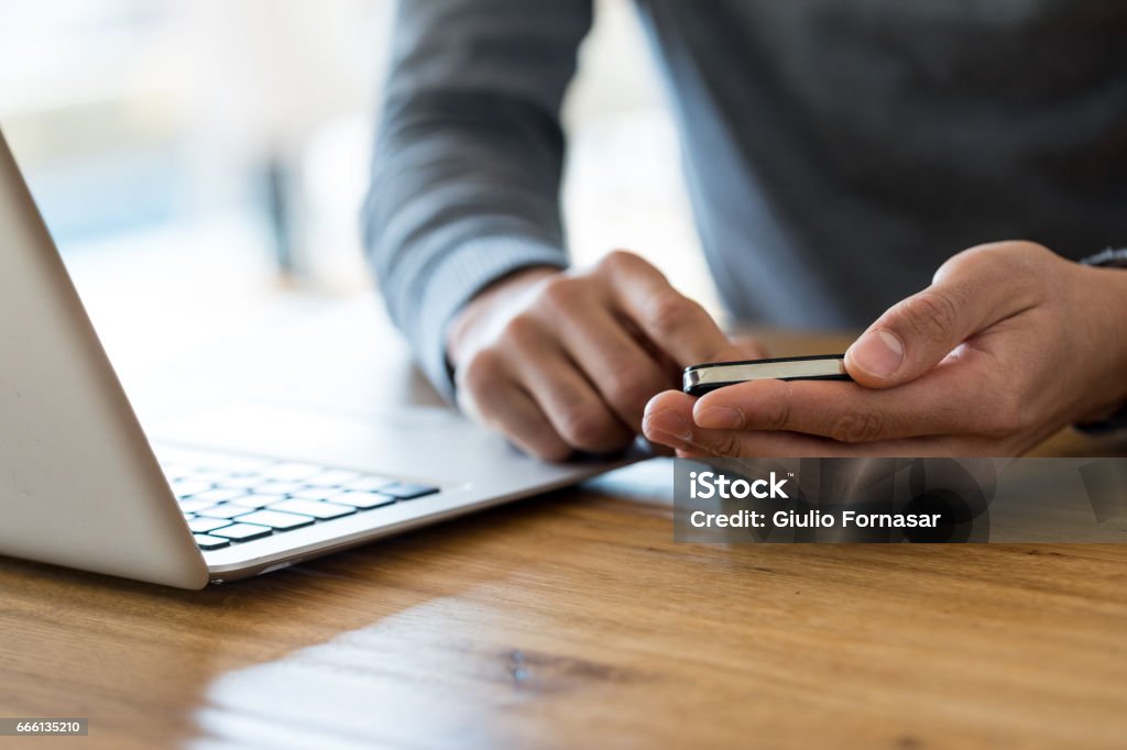 Cropped of man holding cell phone at working place Close-up of unrecognizable man in grey sweater sitting at wooden table next to laptop and holding cell phone in both hands Contact Us Stock Photo