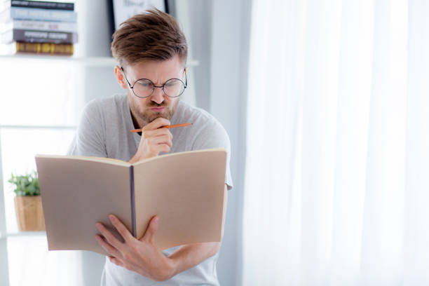 istock Handsome guy in eyeglasses is reading book preparing exam and thinking with standing at the living room - education concept. 666147122