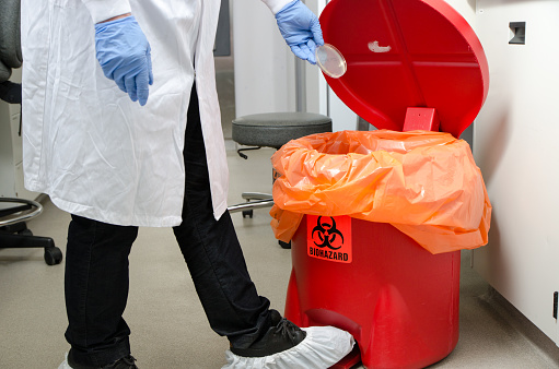 Laboratory worker dropping plastic waste in biohazrd orange trash can

