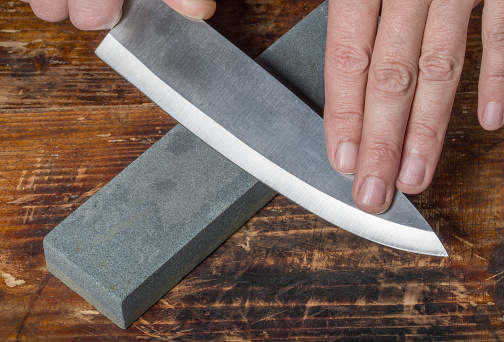 Afilado de cuchillos. Manos sosteniendo cuchillo y piedra de afilar en la vieja tabla de corte de madera. photo