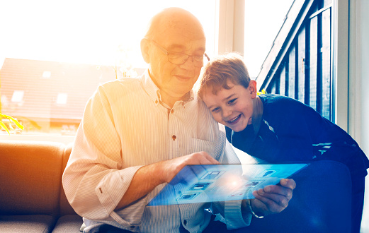 Concept of internet of things, where a digital smart screen controls household items like door locks, washing machine and surveillance camera. The screen looks futuristic and has a see through display. Senior man interacts with the touchscreen. Grandfather and grandson plays with the tablet and enjoy themselves. 