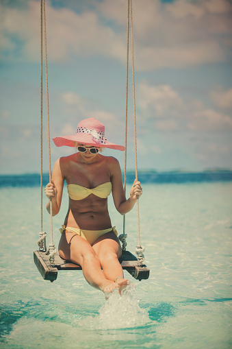 Attractive and sensuality woman in on the swing. She is swinging above water, very close to water so she is crossing through water with legs. With both hands she is holding for swing. On her head she have pink hat with white and black ribbon and sunglasses. She dressed yellow bikini. On the sky there is some clouds but it's sunny day. Water is crystal clear and she is looking down in the water.