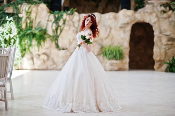 Charming red-haired bride with wedding bouquet at hand posed at great wedding hall with caves and vegetation indoor