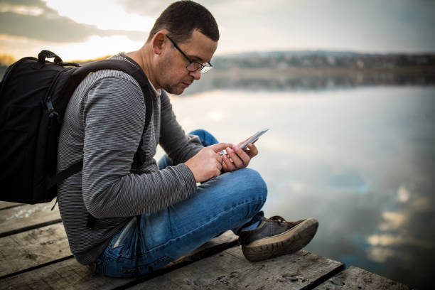 Man with rucksack sitting on lake pier and using mobile phone