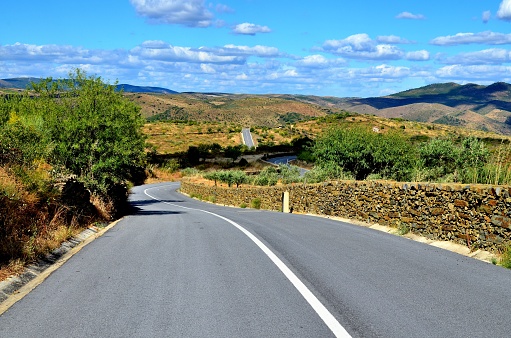 An empty road in Portugal, near the town of Foz Coa, in the Douro valley region. The pictures shows a typically Mediterranean landscape with blue sky, dry hills, olive plantations and stone fences.