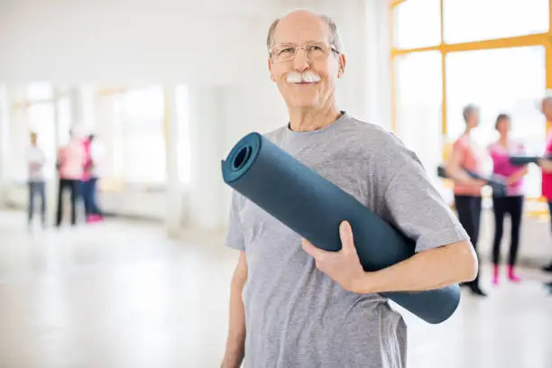 Confident senior man standing in yoga class. Portrait of smiling elderly male holding rolled up exercise mat. He is wearing sports clothing.