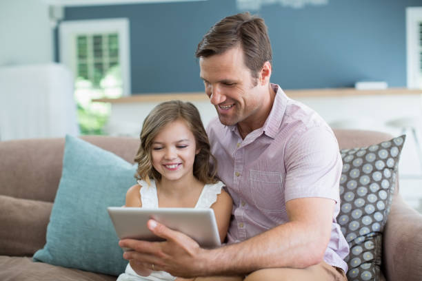 istock Smiling father and daughter sitting on sofa using digital tablet in living room 666473292