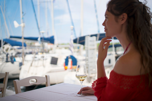 Beautiful caucasian woman sitting at the open air restaurant in marina, drinking wine and enjoying her vacation. Anchored yachts in the back.