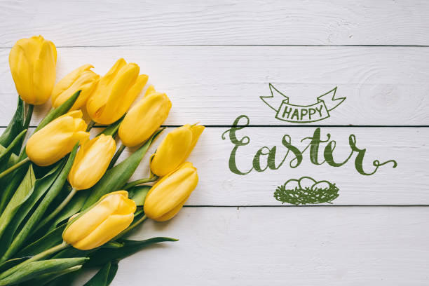 Happy easter hand lettering calligraphy text. Yellow tulips bunch on white wooden planks rustic barn rural table background. Letters, inscription. Beautiful horizontal flat lay postcard.