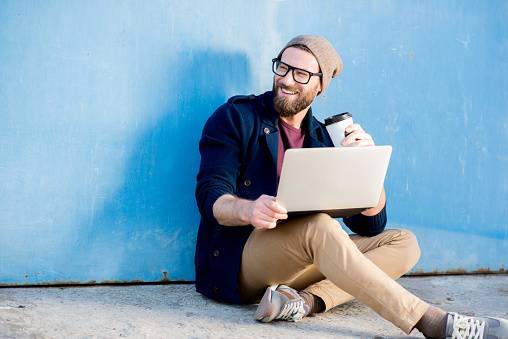 Stylish man dressed casual in sweater and hat working with laptop sitting with coffee near the blue wall background