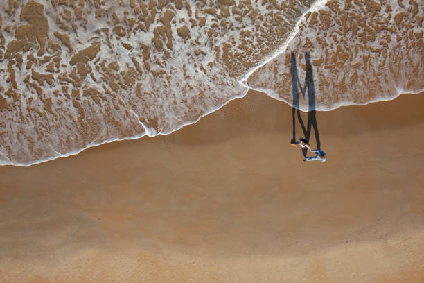Top view of young mixed couple enjoying beach vacation, walking along the water's edge, holding hands. Photo from above of young couple traveling together outdoors on the beachside. Long shadows are clearly visible and emphasizing the bonding.