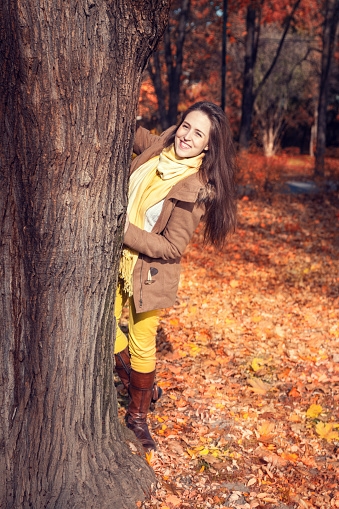 Happy girl peeking out from a tree and having fun in the park. Smiling woman enjoying in autumn day in nature.