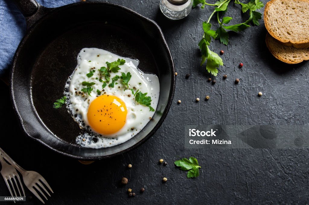 Fried egg Fried egg. Close up view of the fried egg on a frying pan. Salted and spiced fried egg with parsley on cast iron pan and black background. Cast Iron Stock Photo