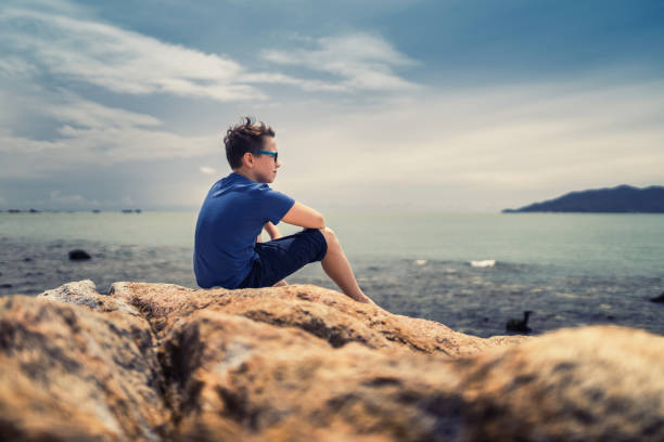Cute boy sitting on rocks and watching at tranquil sea