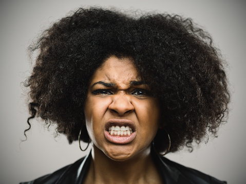 Close-up portrait of sad young afro american woman. Furious female clenching teeth against gray background. Horizontal studio photography from a DSLR camera. Sharp focus on eyes.