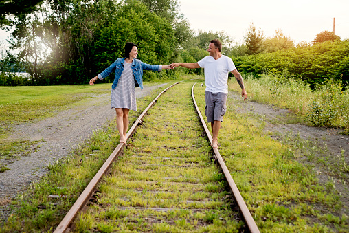 Loving couple trying to balance their way on a railroad track, at the end of a sunny day in summer. They are looking at each other, holding hands. Horizontal full length outdoors shot with copy space. This was taken in Quebec, Canada.