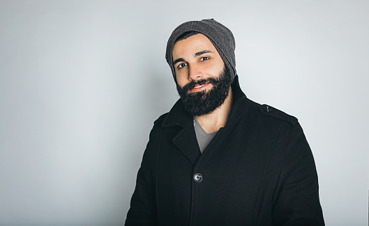 Studio portrait of smiling Indian young hipster bearded male with cap and black coat over white background.