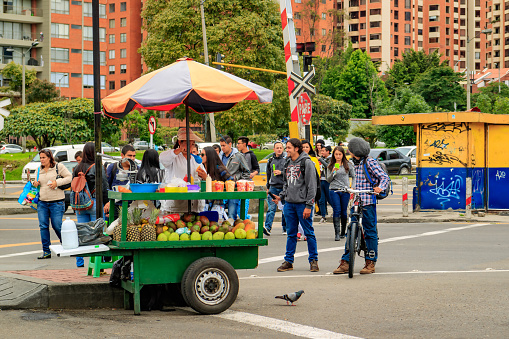 Bogotá, Colombia - March 10, 2017: A Colombian woman sells fresh fruit salads on the intersection of Carrera Novena with Calle 134. She has mangoes, pears, papayas and pineapples on her cart. In the background are people crossing the Carrera and in the far background are some apartment blocks. Photo shot on an overcast morning; horizontal format.