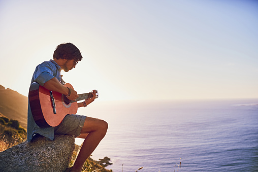 Shot of a young man playing the guitar while sitting at a viewpoint outside