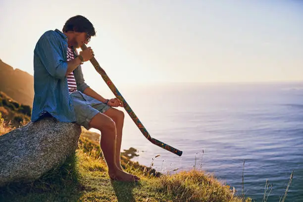 Shot of a young man playing the didgeridoo while sitting at a viewpoint outside