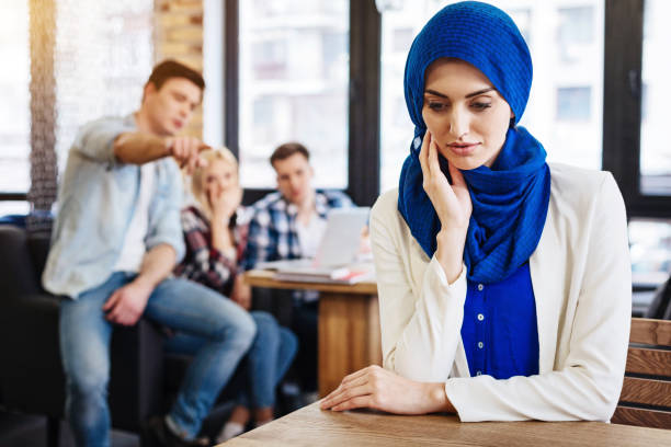 Cheerless muslim woman feeling unjustice from the society Multicultural society. Pleasant beautiful young muslim woman sitting at the table in the cafe while her mates abusing her modest clothing stock pictures, royalty-free photos & images