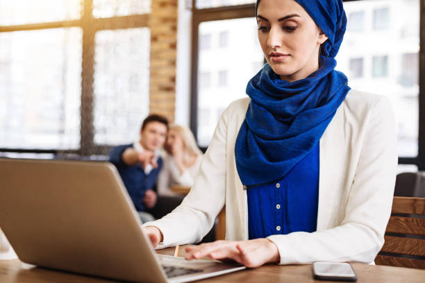 Pleasant muslim businesswoman working on the laptop Involved in work. Pleasant attractive muslim businesswowoman working on the laptop and sitting in the cafe while young couple spreading gossips behind her back racism stock pictures, royalty-free photos & images