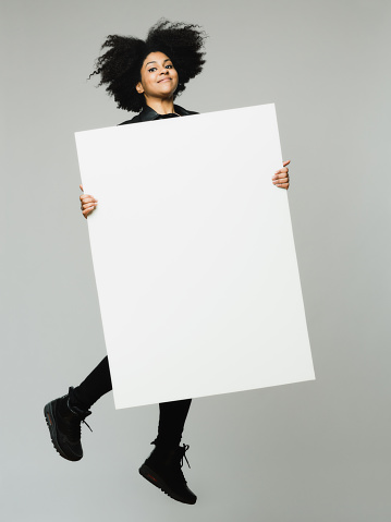 Full length portrait of cheerful young african woman jumping and holding a blank billboard. Vertical shot of real black woman with empty poster over gray background. Studio photography from a DSLR camera. Sharp focus on eyes.